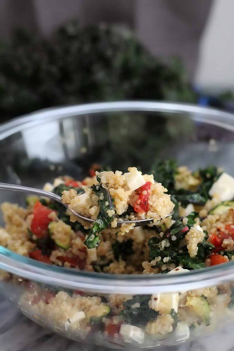 A spoon takes a scoop of quinoa, vegetables, and feta cheese from a large glass mixing bowl, with a bunch of kale in the background.