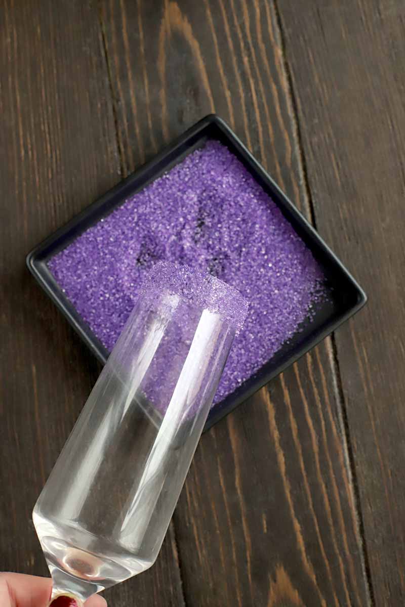 Closeup overhead shot of fingers with red nail polish holding the stem of a champagne flute and dipping the rim into a square, shallow dish of purple sanding sugar, on a dark brown wood background.