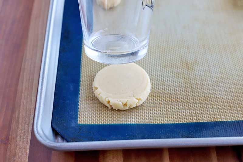 A pint glass being used to flatten round balls of dough, on a blue and tan silicone pan liner set into a rimmed baking sheet, on a striped wood surface.