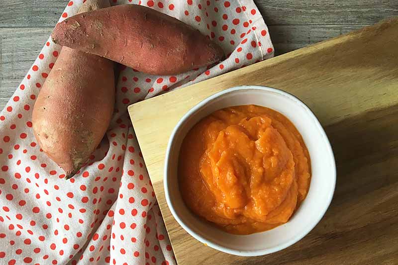 Horizontal image of a white bowl with mashed sweet potatoes on a wooden cutting board.