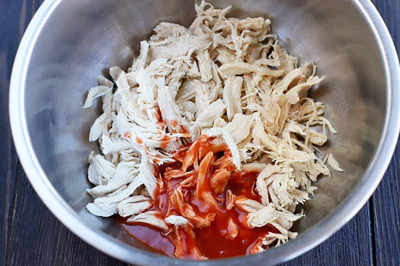 A stainless steel mixing bowl of shredded cooked chicken breast and red hot sauce, on a dark brown wood surface.