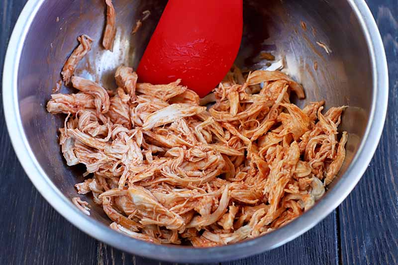 Shredded chicken mixed with hot sauce in a stainless steel mixing bowl with a red spatula, on a dark brown wood surface.