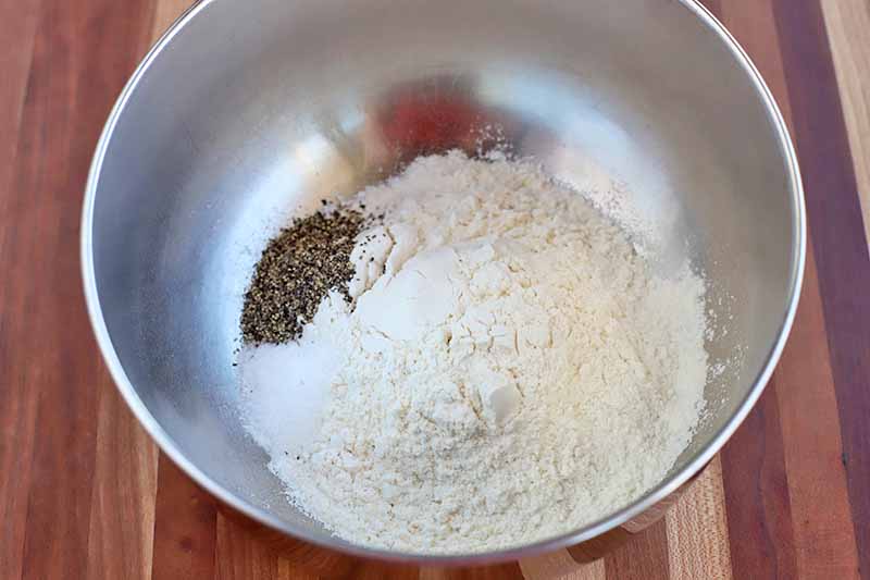 Salt, black pepper, and flour for a dredging mixture in the bottom of a stainless steel mixing bowl, on a brown striped wood table.