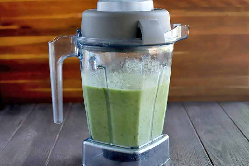 A pitcher-style blender is filled with a green smoothie mixture, on a dark brown wood surface with a lighter brown background.