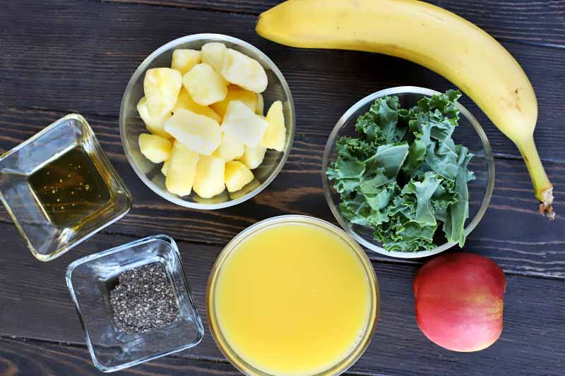 Overhead shot of a banana and an apple, and small round and square glass bowls of torn green kale leaves, orange juice, frozen pineapple chunks, chia seeds, and honey, on a dark brown wood table.