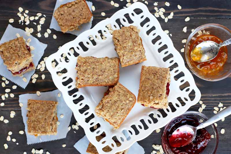 Homemade breakfast bars on a white serving dish and squares of parchment paper, with two bowls of red and orange jelly with spoons to the right, and scattered uncooked oats, on a dark brown wood surface.
