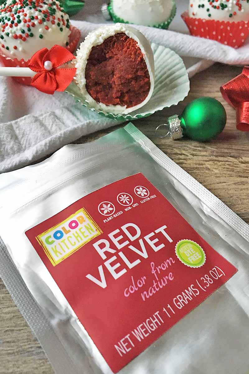 Vertical image of Color Kitchen's Red Velvet pigment in front of a half-eaten dessert pop with festive decorations.
