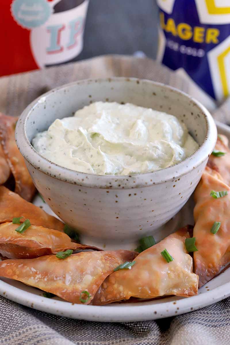 A small bowl of blue cheese dip on top of a round platter of chicken wonton bites with chopped green onions on top, with cans of beer in the background.
