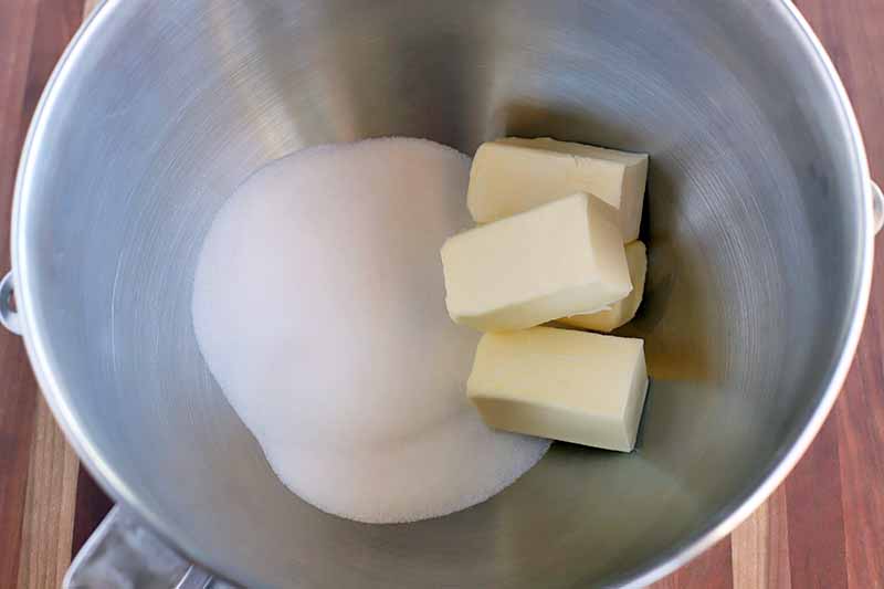 Sticks of butter and sugar in the bottom of a stainless steel stand mixer bowl, on a wood surface.