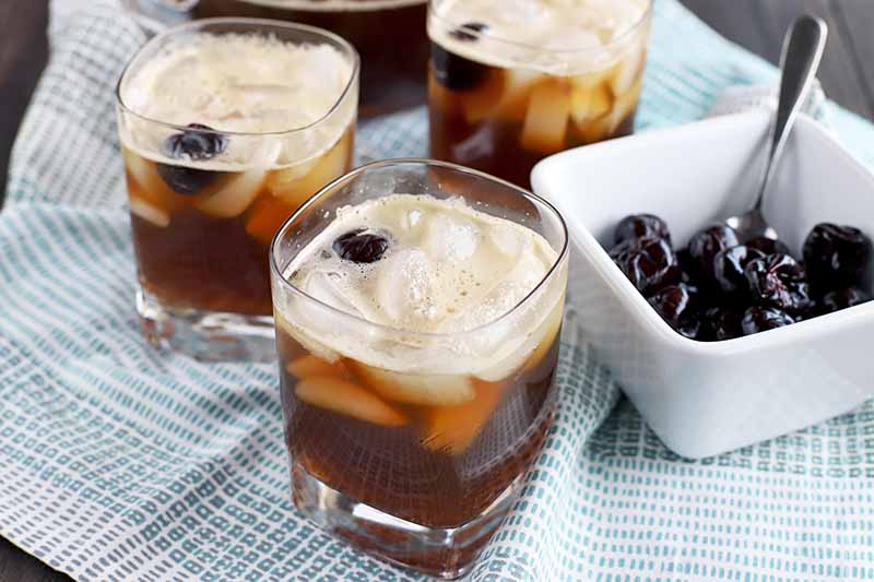 Three square lowball glasses of cherry cola beer punch garnished with maraschino cherries, with more of the preserver fruit in a small square dish to the right with a spoon, on a blue and white cloth on top of a dark brown wood surface.