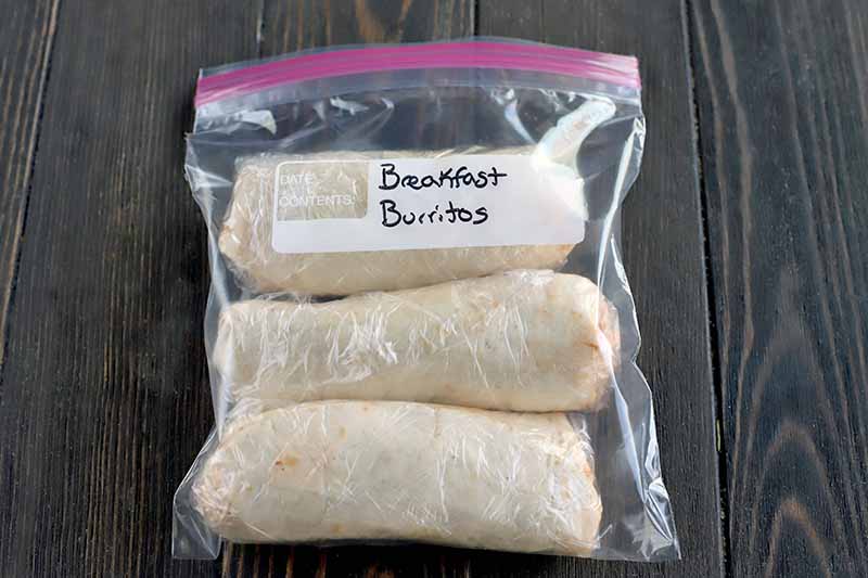 Three burritos wrapped in plastic and placed in a zip-top freezer bag labeled in black permanent market, on a dark brown wood table.
