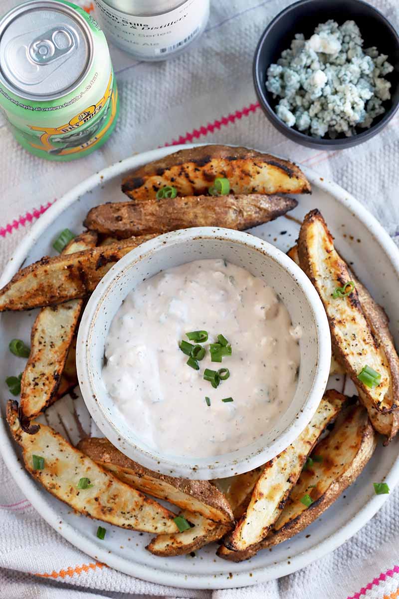 Vertical image of a plate of potato wedges surrounded a bowl of a white creamy dip.