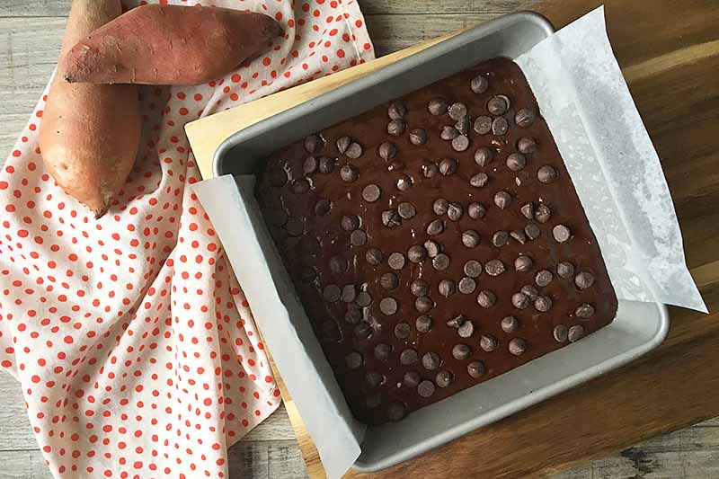 Horizontal image of an unbaked brownie mixture topped with chocolate chips on a wooden cutting board.