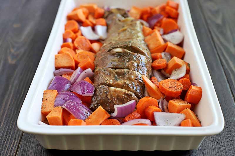 Roasted pork tenderloin surrounded by orange root vegetables and purple onion in a square ceramic baking dish, on a dark brown wood table.