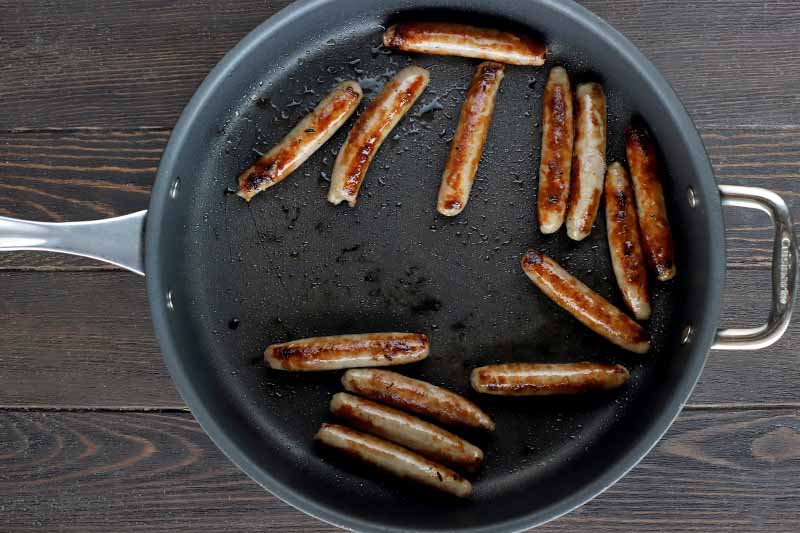 Sausage links are browning in a large nonstick skillet, on a dark brown wood surface.
