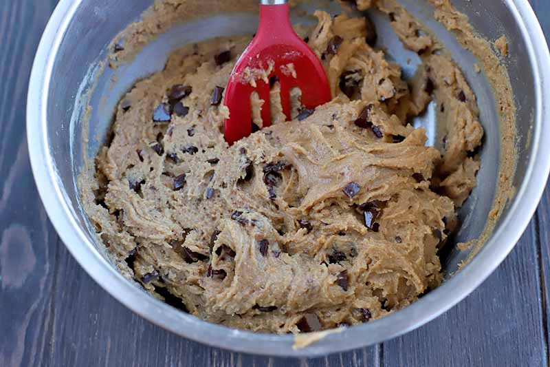 A red slotted rubber utensil stirs a dough mixture studded with dried cherries and chocolate chunks in a stainless steel mixing bowl, on a dark brown wood surface.