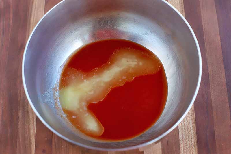 Hot sauce and melted butter at the bottom of a stainless steel bowl, on a striped wood surface.