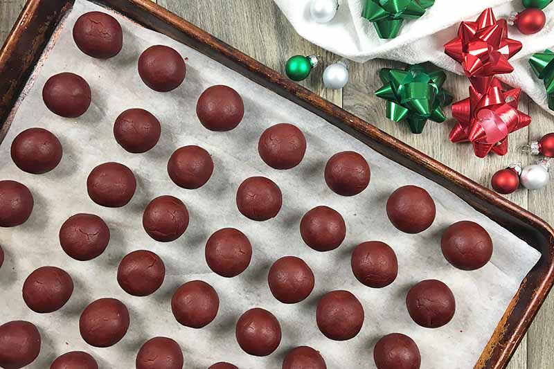 Horizontal image of a sheet pan lined with parchment paper, with rows of red velvet cake balls next to holiday decorations.