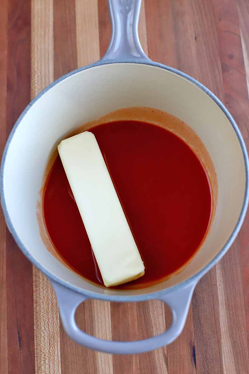 Overhead shot of a light blue and cream-colored enameled saucepan with vibrant red hot sauce and a stick of butter at the bottom, on a striped wood surface.
