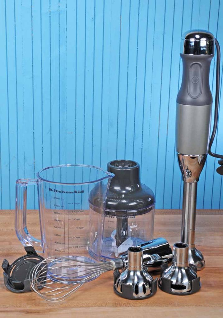 The KitchenAid KHB2561 5-speed Hand Blender Set with attachments on maple butcher block table with a blue background.