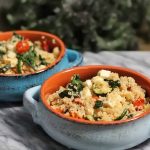 Two terra cotta handled bowls with a light blue and darker blue glaze, filled with a mixture of quinoa, vegetables, and feta, on a marble surface with a bunch of kale in soft focus in the background.