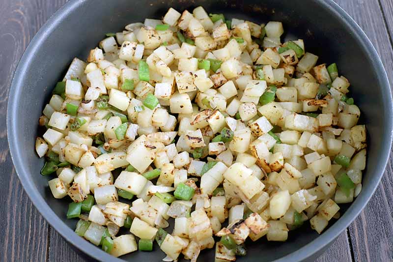 Diced green bell pepper, onion, and potato are being prepared in a large nonstick frying pan, on a dark brown wood surface.