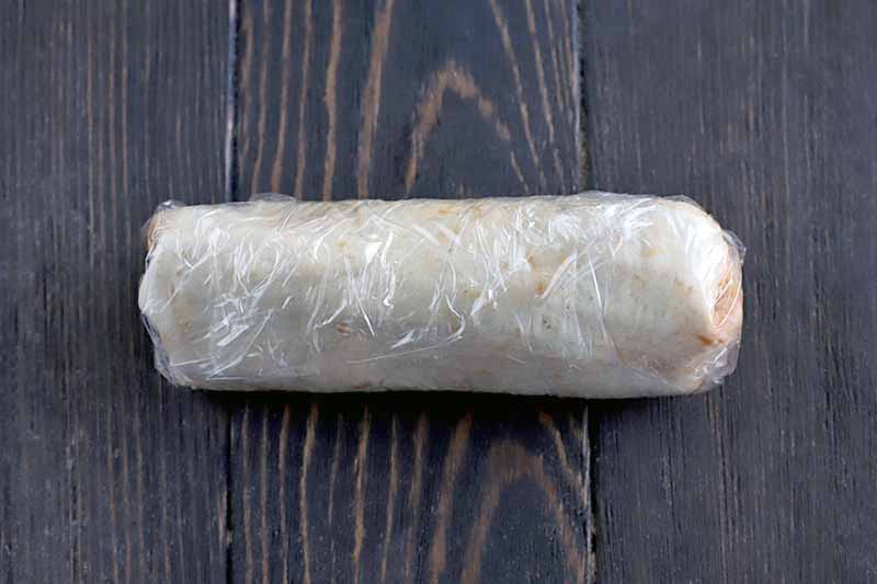 A burrito wrapped in plastic on a dark brown wood table.