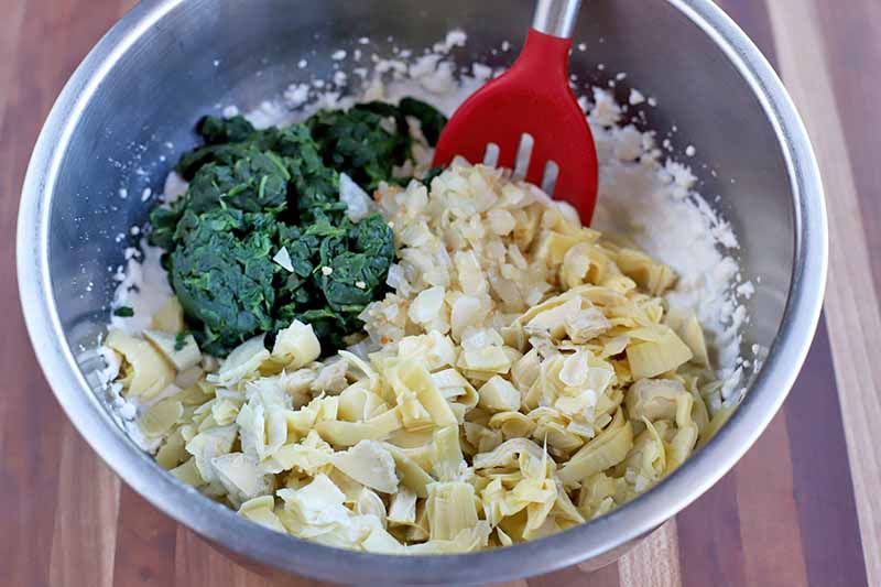 A slotted red silicone spoon with a silver metal handle is being used to stir a mixture of mayonnaise, chopped artichoke hearts, and drained frozen spinach, on a large stainless steel mixing bowl on a striped wood surface.