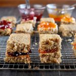 Gluten-free oat sandwich bars filled with orange and red fruit jam on a metal cooling rack, with two small glass bowls of jam with spoons stuck into them in shallow focus in the background, on a dark brown wood table with a brown background.