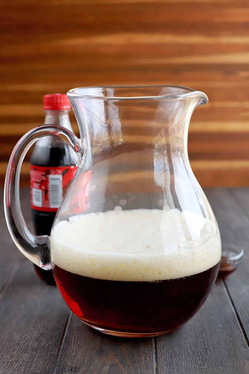 A glass pitcher is filled about halfway with a frothy brown liquid, with a bottle of soda in the background, on a dark brown wood surface with a striped brown backdrop.