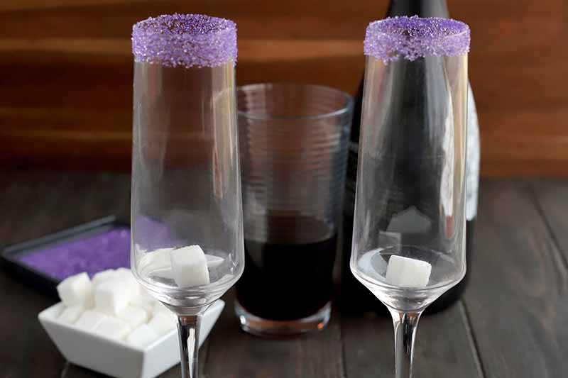 Closely cropped image of two champagne flutes with white sugar cubes at the bottom and purple rims, with more of both garnishes in small dishes in the background beside a pint glass containing a brown liquid and a bottle of champagne, on a dark brown wood table in front of a striped brown backdrop.