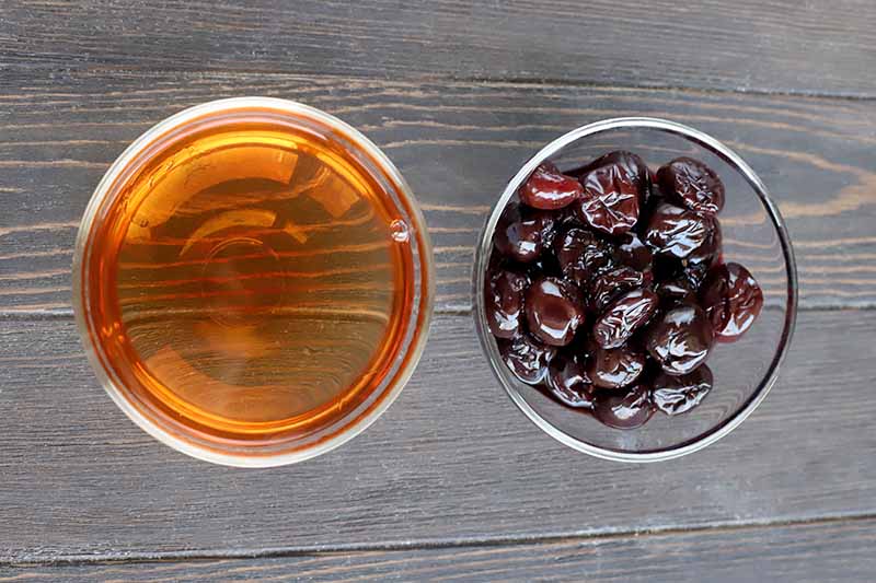 Overhead shot of two small glass bowls of sherry and maraschino cherries, on a dark brown wood surface.
