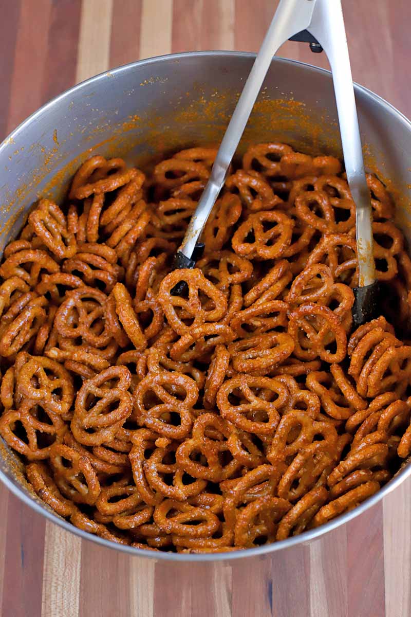 Closely cropped overhead shot of a stainless steel mixing bowl of pretzels coated in sauce, with a pair of metal tongs, on a striped wood surface.