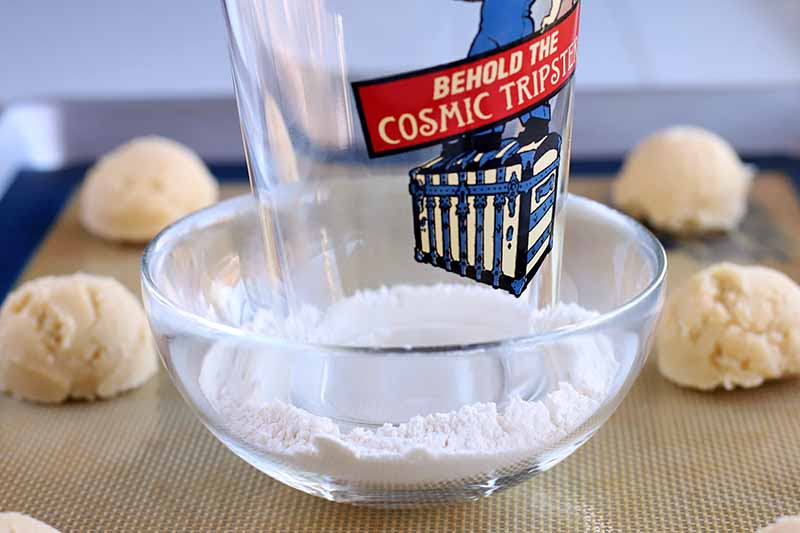 A pint glass rests in a small glass bowl of flour, on top of a silicone pan liner with portioned dough arranged around it, set into a rimmed baking sheet.