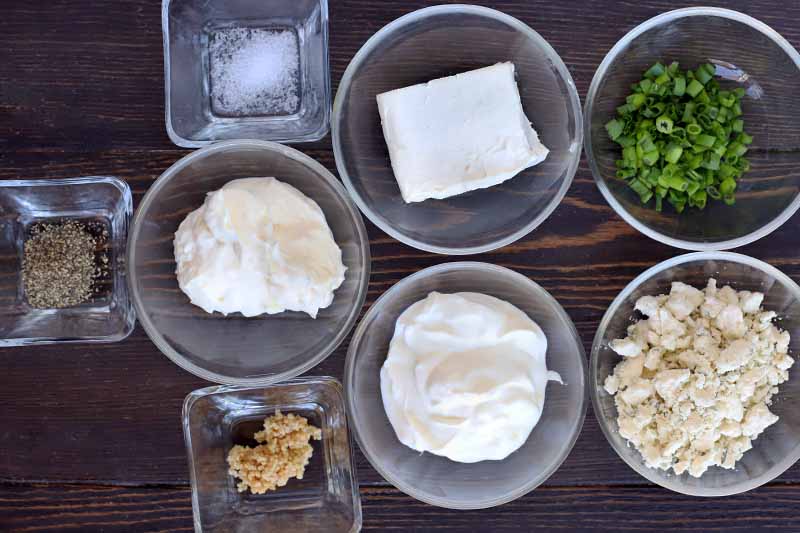 Overhead shot of square and round glass bowls of salt, pepper, grated cheese, cream cheese, sour cream, blue cheese, garlic, and scallions, on a dark brown wood surface.
