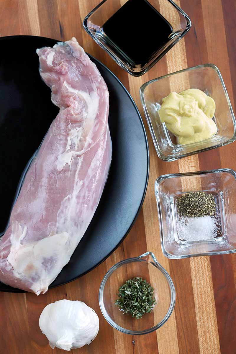 Overhead shot of a raw pork tenderloin on a black plate, surrounded by small round and square glass dishes of balsamic vinegar, garlic, spices, herbs, and garlic, on a striped wood surface.