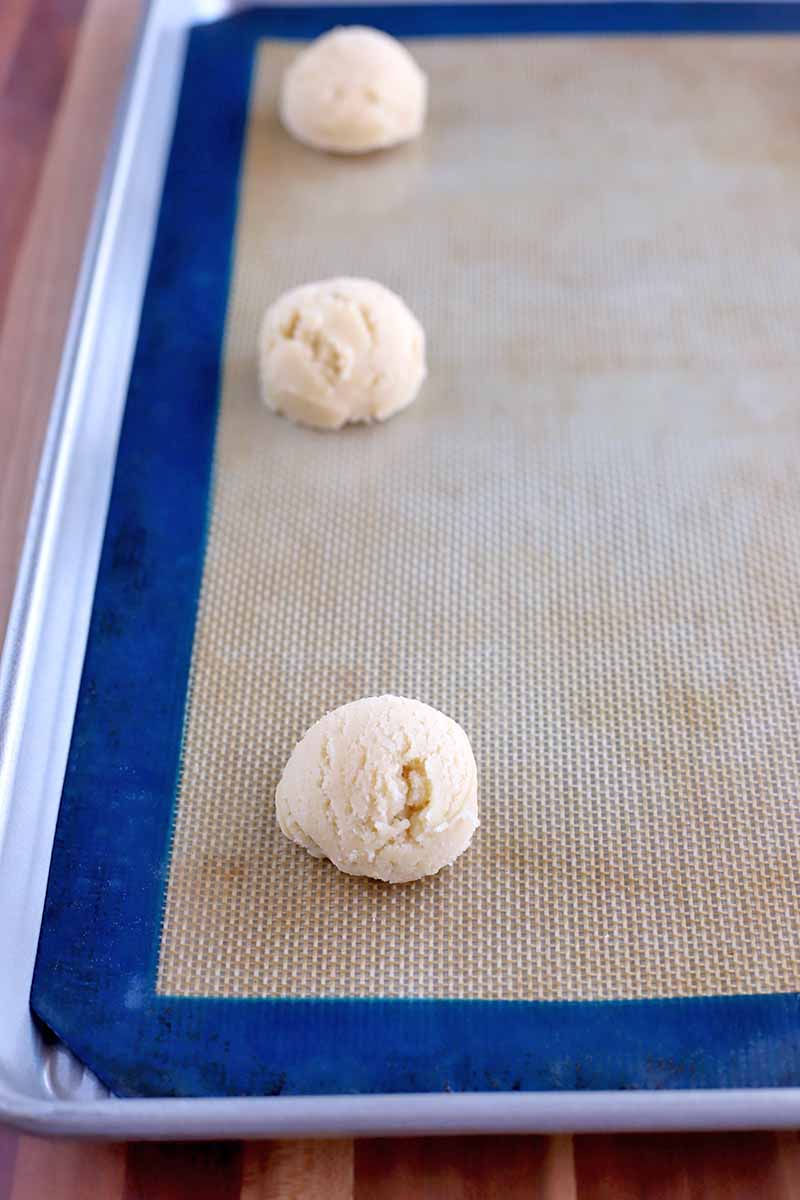Three portioned balls of dough on a blue and beige silicone pan liner set into a rimmed baking sheet, on a wood surface.