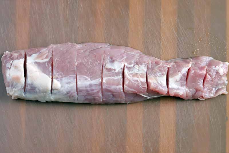 A raw pork tenderloin with slits cut into the surface at even intervals, on a clear translucent plastic cutting board on top of a wood surface.
