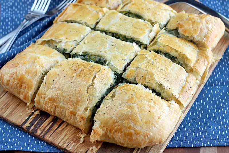 Twelve slices of spinach in puff pastry arranged in rows on a wood serving board and shot at an oblique angle, with forks on top of a blue cloth with white flecks, on a wood background.