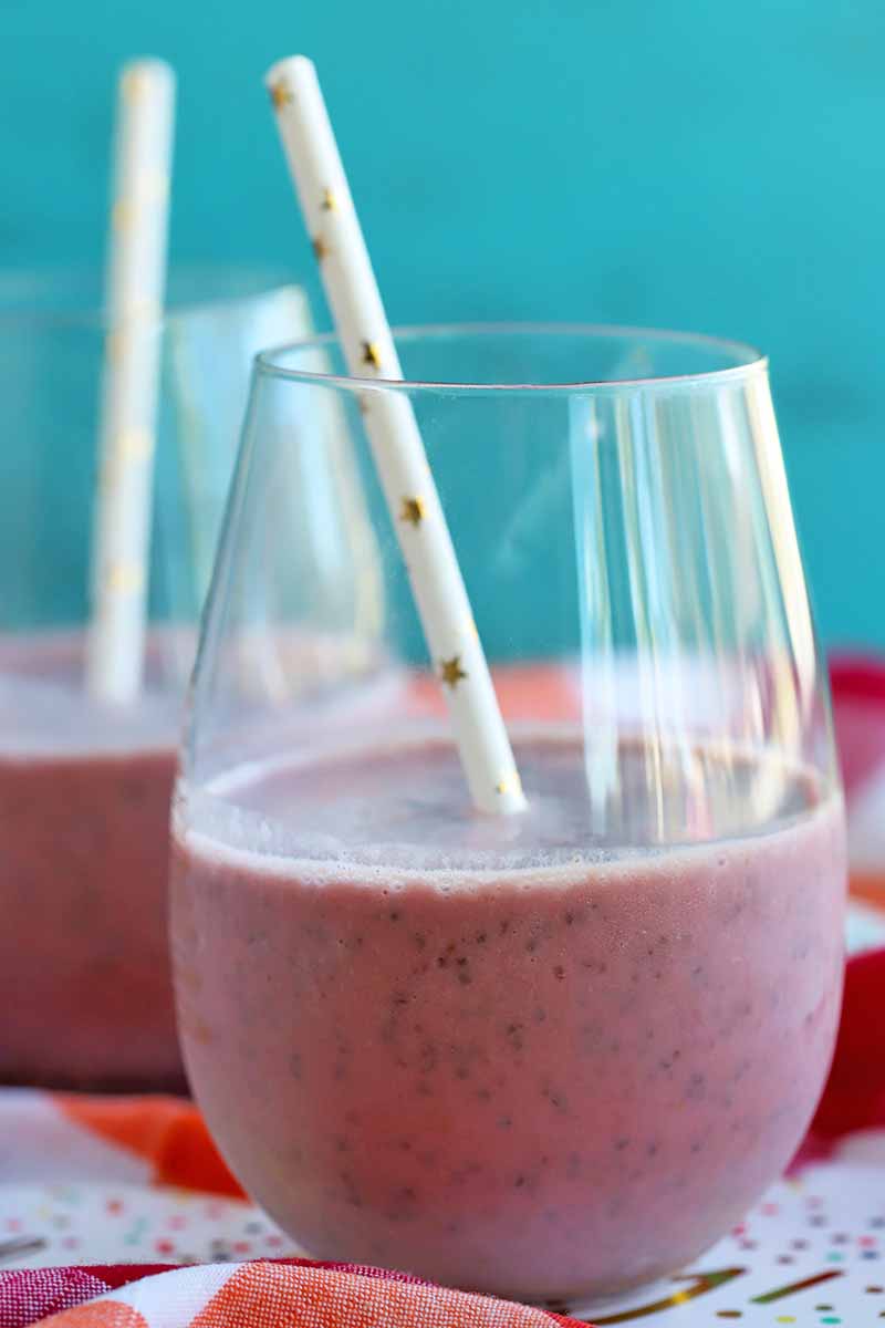 Vertical image of two clear stemless wine glasses filled halfway with a pink fruit smoothie, with white paper straws decorated with gold metallic stars, on a red and white checkered cloth with a robin's egg blue background.