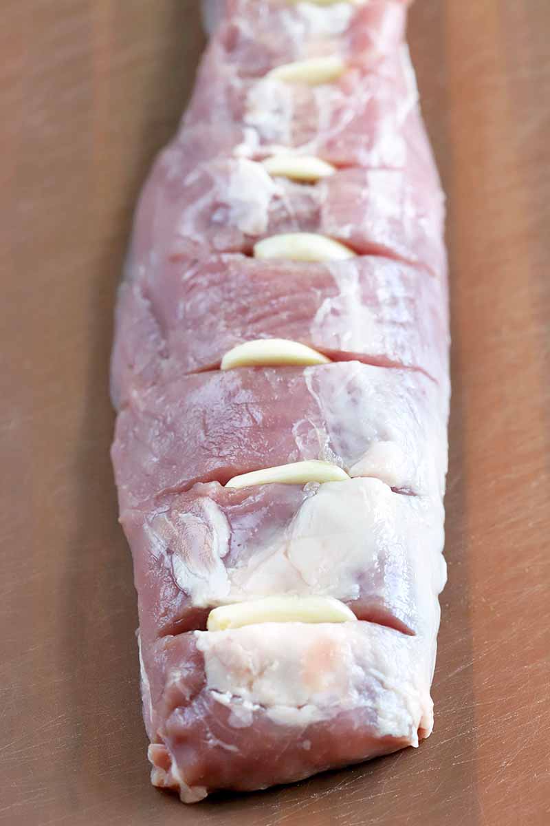 Overhead vertical shot of a raw pork tenderloin with slits cut in the top and stuffed with thin slices of garlic, on a brown surface.