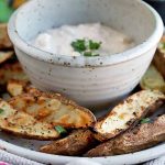 Horizontal image of a bowl of a creamy condiment on a plate with grilled spuds.