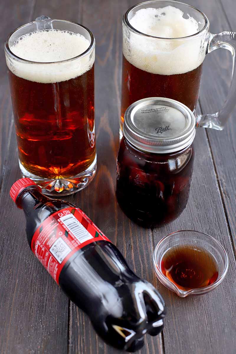 Vertical image of two glass mugs filled with beer, a glass jar with a metal lid filled with maraschino cherries and sherry, a small glass bowl of cherry liqueur, and a small plastic bottle of cola with a red label and lid, resting on its side on a dark brown wood surface.