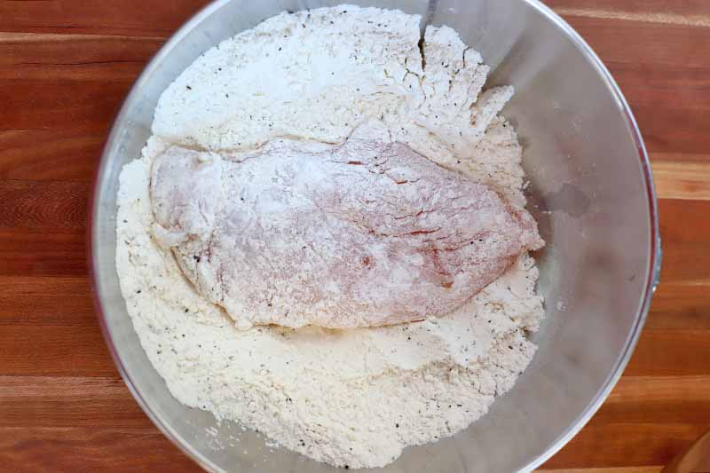 A piece of raw chicken breast is being dredged in flour in a stainless steel bowl, on a brown wood surface.