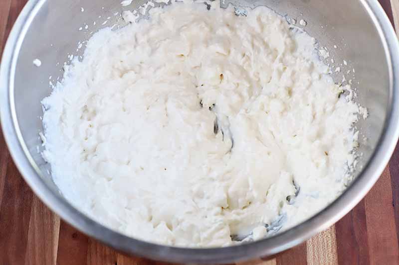 A mayonnaise and sour cream mixture for making dip in a large stainless steel mixing bowl, on a striped brown wood background.