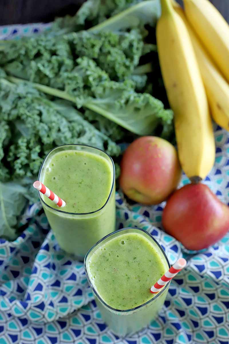 Oblique vertical shot of apples, bananas, kale, and two green smoothies with red and white striped straws, on a dark and light blue cloth with a triangular pattern.