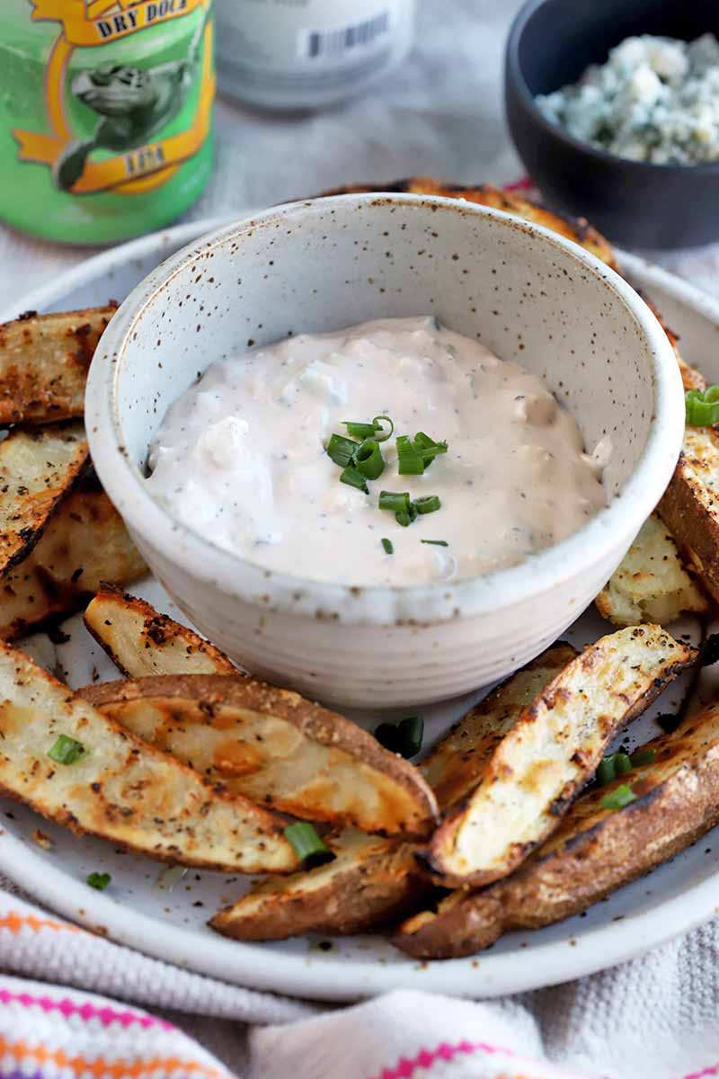 Vertical image of a bowl of white dip with green garnish and a plate of potato wedges.