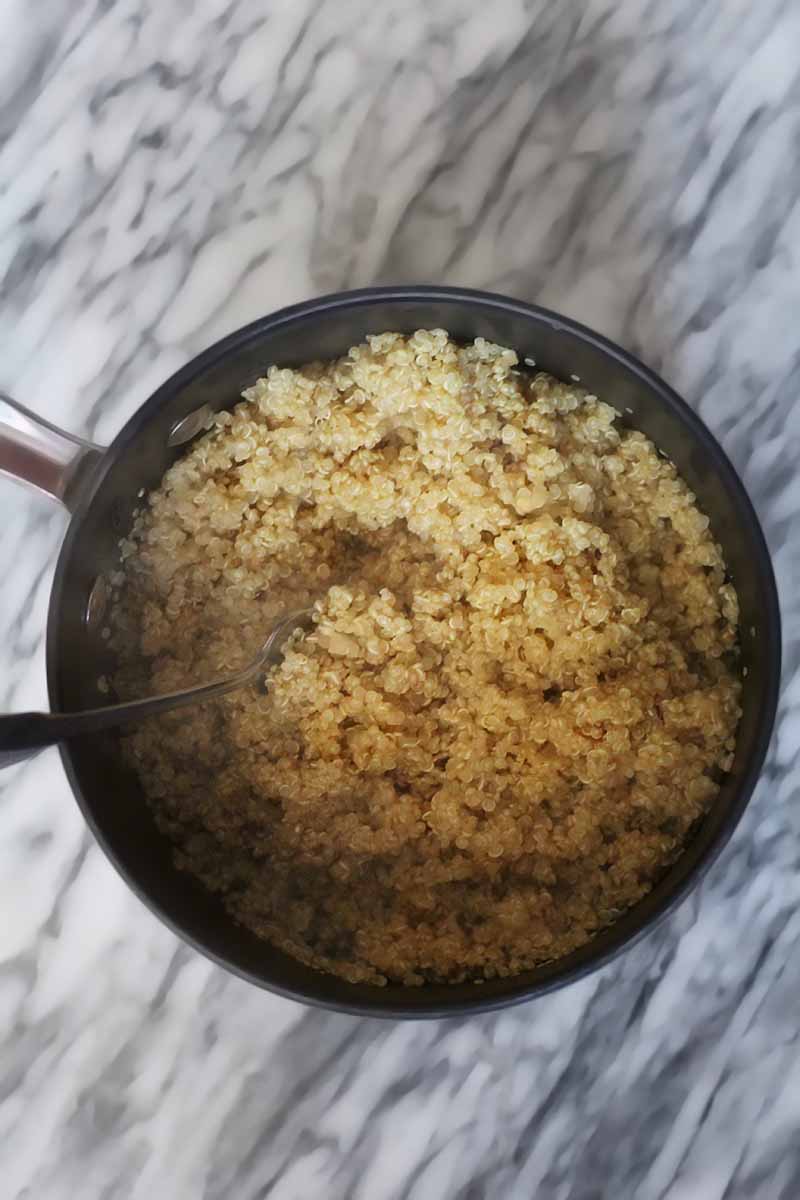 Overhead shot of steaming cooked quinoa in a saucepot with a spoon, on a marble surface.