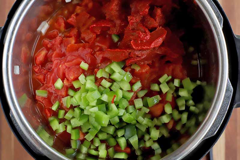 Closeup closely cropped overhead shot of a stainless steel and black plastic slow cooker filled with red canned tomatoes and tomato paste, and diced green bell pepper.