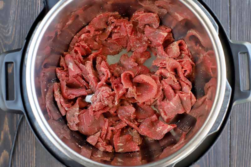 Thinly sliced raw beef in the bottom of a slow cooker, on a dark brown wood surface.
